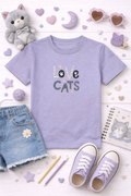 Purple t-shirt with 'Love Cats' text, denim shorts, lavender sneakers, and cat-themed accessories on a light background.